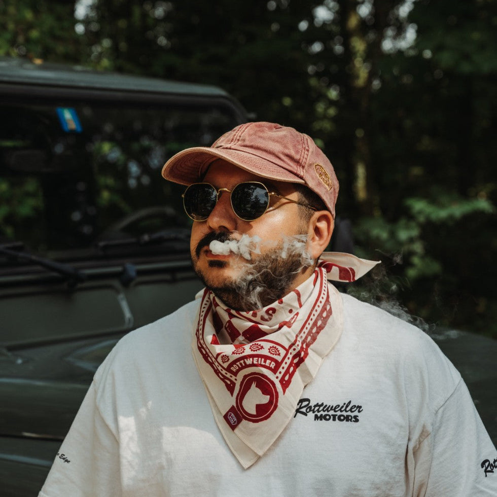 Rider wearing the red and white Rottweiler Motors bandana beside a green Defender with smoke drifting across the portrait.